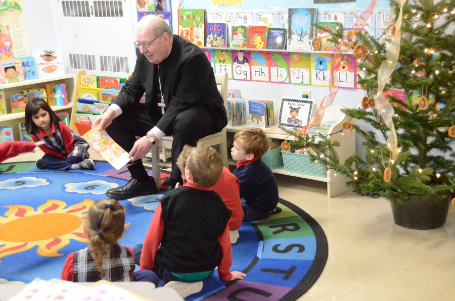 The bishop is seating in a chair with children surrounding him as he reads a book.