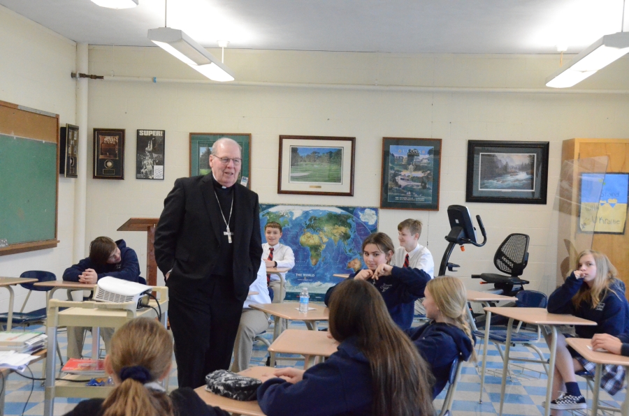 The bishop standing in the middle of desks with students.